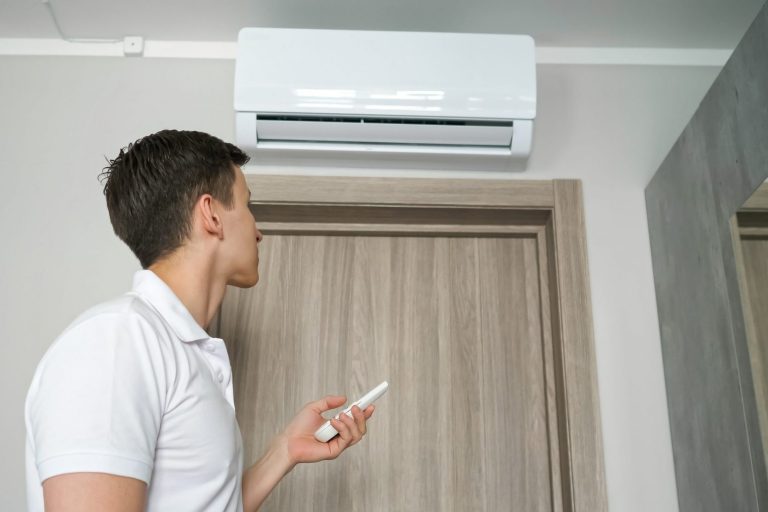 Photo of man looking at AC unit with remote control in hand illustrates blog: "My Air Conditioner Won’t Stay On. What To Do?"