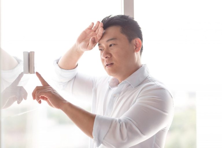 Photo of sweaty man looking at thermostat illustrates blog: "Why Is My Thermostat Changing on Its Own?"